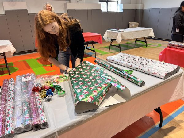 Junior Lucia Butterfield wraps a gift at her wrapping station with all the gift wrapping essentials: various rolls of paper, bows, tape, scissors, and name tags. Most students worked their own table, wrapping many different gifts, while others sat with kids and babies of the parents picking out gifts. The students volunteered at Neighbors Link Yonkers location at Dayspring Community Center.